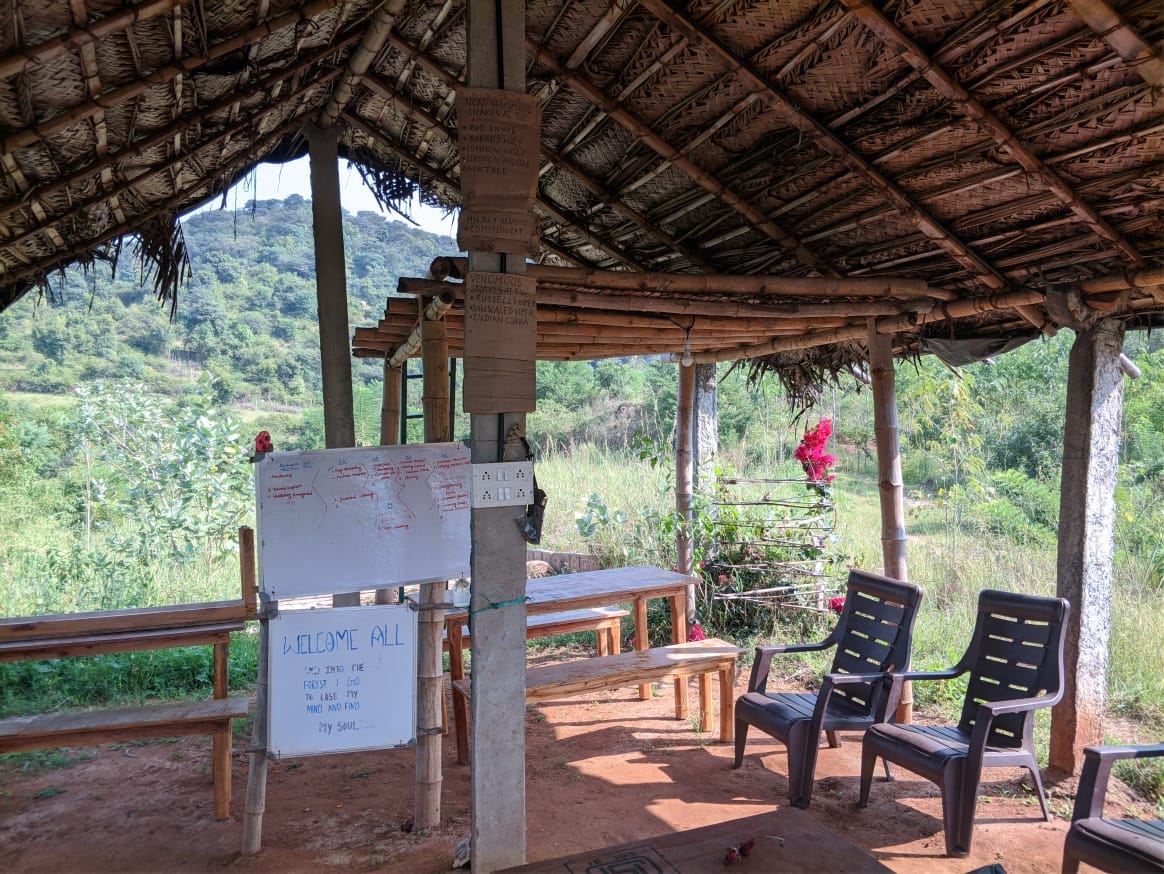 Thatched bamboo pavilion at TVC overlooking the Melagiri hills, set up with benches and a whiteboard for a camp-night session.