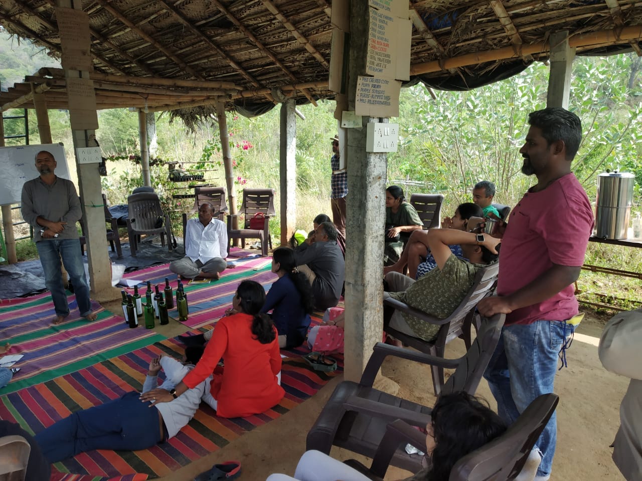 Workshop participants seated on colorful mats under TVC's bamboo pavilion, listening to a facilitator.